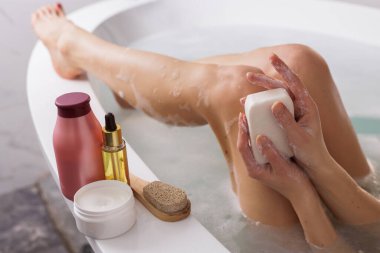 Yanında köpük, vücut ve saç bakım ürünleri olan bir küvette otururken elinde sabun tutan bir kadın.