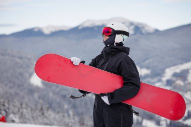 Siyah kıyafetli bir snowboardcu elinde kırmızı bir snowboard ile karlı bir dağda manzaralı bir manzarayla.