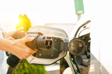Bir istasyonda şarj olan elektrikli bir araç, şarj portuna yakın çekim. Arka planda, başka bir araba park edilmiş, ve yeşil bir çalı görünür, güneş ışığıyla aydınlatılmış..