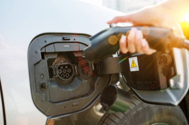 Bir istasyonda şarj olan elektrikli bir araç, şarj portuna yakın çekim. Arka planda, başka bir araba park edilmiş, ve yeşil bir çalı görünür, güneş ışığıyla aydınlatılmış..
