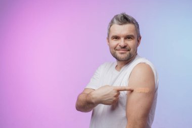 Mutlu yetişkin Hintli İspanyol adam pembe mavi arka planda izole edilmiş aşılardan sonra kolunda yama gösteriyor. modern aşılama ve insanların aşılanması