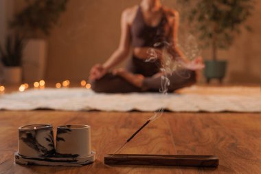 Tütsü ve mumlar odak noktasında yanıyor. Arka planda meditasyon yapan bir kadın, sakin bir ortam yaratıyor..