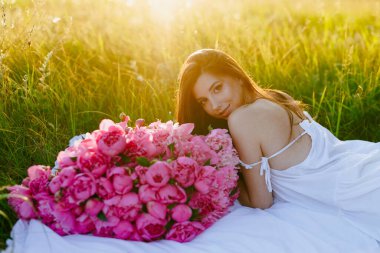 Geniş bir buket pembe şakayıklı ve beyaz elbiseli genç bir kadın sıcak güneşin ışınlarında bir ağacın üzerinde bir battaniyenin üzerinde yatıyor. Reklam, sosyal medya, yaşam tarzı blogları ve romantik kavramlar için ideal.