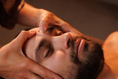 Sakallı bir adamın kaplıcada rahatlatıcı bir yüz masajı yaptırdığı yakın plan. Ilık aydınlatma, bu kişisel bakım ve sağlık tedavisinin barışçıl ve tedavi edici atmosferini arttırır.