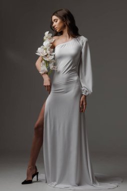 Beyaz orkideler tutan tek omuzlu beyaz elbiseli zarif bir kadın. Minimalist stüdyo ortamında zarafet, kadınlık ve sofistike bir ortam..