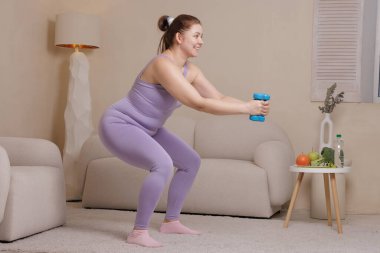 Spor kıyafetleriyle gülümseyen, kilolu bir kadın evinde dambıllarıyla çömelmiş, rahat iç mekanlarda spor, kilo kaybı ve sağlıklı yaşam tarzını destekliyor..