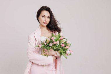 Smiling woman in a pink suit holding a bouquet of fresh tulips, posing against a neutral background in a soft, feminine, and elegant spring-inspired look clipart