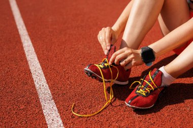 Bir bayan sporcu bir stadyumda spor ayakkabısının bağcıklarını bağlayıp yarışa hazırlanıyor. Ellerinin yakın çekimi ve güneş ışığındaki profesyonel çıkıntılar.