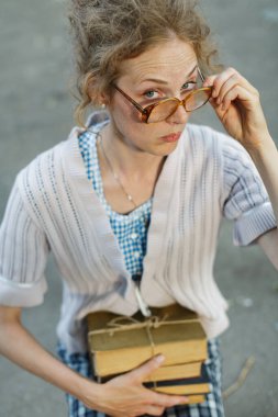 Büyük gözlüklü inek genç bir kadın güneş ışığında ayakta dururken ağır kitaplar taşımaya, esnemeye ya da nefes nefese kalmaya çalışıyor..