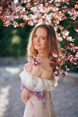 Çiçek açan kiraz çiçeklerinin arasında gülümseyen genç bir kadın. Dalgalı kollu çiçekli bir elbise giyer, güneşli bir bahçede neşeli ve huzurlu bir hava yayar..