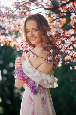 Çiçek açan kiraz çiçeklerinin arasında gülümseyen genç bir kadın. Dalgalı kollu çiçekli bir elbise giyer, güneşli bir bahçede neşeli ve huzurlu bir hava yayar..