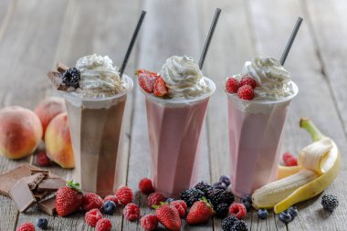Üç lezzetli milkshake, çırpılmış krema, taze böğürtlen, çikolata ve meyve, kırsal bir ahşap masada uzun bardaklarda servis edilir..