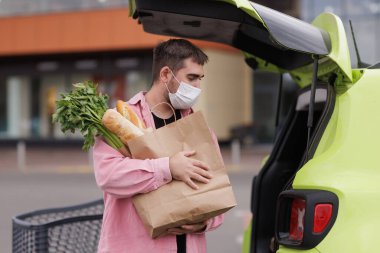 Yüz maskesi takan bir adam marketin önünde içi taze yiyecek, ekmek ve yeşillik dolu bir poşet tutan bir arabaya yiyecek yükler..