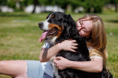 Genç bir kadın güneşli bir parkta mutlu Bernese Mountain Dog 'una sarılıyor, dışarıda sıcak bir aşk ve arkadaşlığın tadını çıkarıyor.
