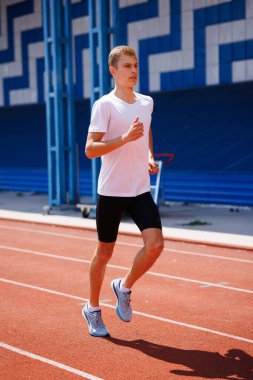 Genç erkek atlet kırmızı bir pistte koşuyor, spor kıyafetleri giyiyor, güneşli bir günde açık havada antrenman yapıyor, hız ve performansa odaklanıyor..