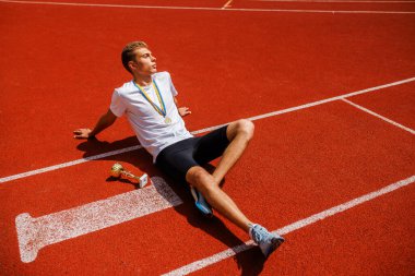 Yarışı kazandıktan sonra koşu pistinde dinlenen yorgun genç bir sporcu, yanında bir kupa, başarıyı ve sıkı çalışmayı simgeleyen bir madalya takıyor..