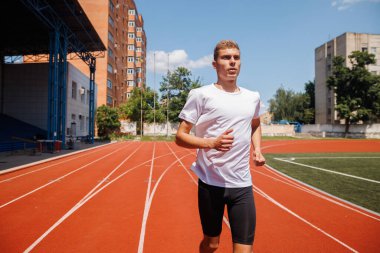 Genç erkek atlet kırmızı bir pistte koşuyor, spor kıyafetleri giyiyor, güneşli bir günde açık havada antrenman yapıyor, hız ve performansa odaklanıyor..