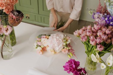 Kadın çiçekçi, cam vazolarda renkli çiçeklerle çevrili çiçek atölyesinde taze çiçeklerle narin bir buket oluşturuyor. Çiçekçi