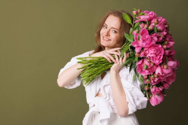 Beyazlı bir kadının zeytin yeşili arka planda tuttuğu büyük bir buket pembe şakayık.