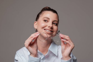Dişlerinde yuvarlak gözlüklerle gülümseyen genç bir kadın, kendine güvenen ve mutlu bir ifade, beyaz gömlek içinde şık, gri arka planda modern bir portre..