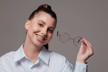 Dişlerinde yuvarlak gözlüklerle gülümseyen genç bir kadın, kendine güvenen ve mutlu bir ifade, beyaz gömlek içinde şık, gri arka planda modern bir portre..