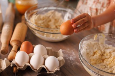 Çocuk sıcak bir mutfakta un, hamur, oklavalar ve karıştırma kaseleriyle yumurta tutarken.