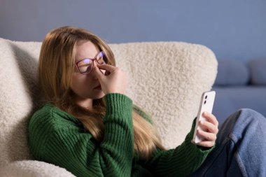 Gözlüklü genç bir kadın akıllı telefon kullanırken gözlerini ovalıyor. Evde dijital yorgunluk ve göz yorgunluğu hissediyor..