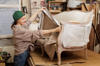 Gülümseyen kadın döşemeci atölyedeki mobilyaları restore ederken kumaşı eski kanepe çerçevesine geriyor..