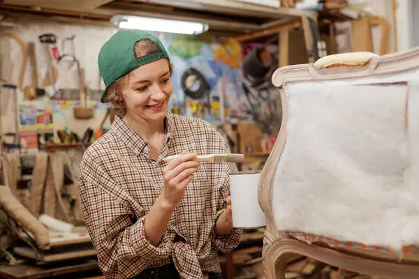 Gülümseyen kadın zanaatkar atölyedeki döşeme yenilenmesi sırasında sandalyenin çerçevesine boya sürüyor..