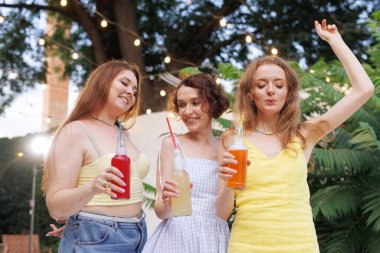 Üç neşeli genç kadın, arka planda ışıklar olan açık hava yaz partisinde renkli şişelenmiş içkiler içiyorlardı..
