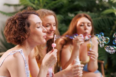 Dışarıda sabun köpüğü üfleyerek eğlenceli ve kaygısız bir yaz gününün tadını çıkaran üç neşeli genç kadın..