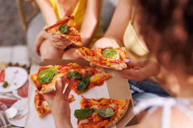 Arkadaşların açık havada pizza paylaştığı en iyi manzara, bir kutunun içinde peynirli dilimlere uzanan ellerin yakın çekimi..