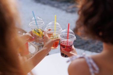 Bir grup arkadaş yaz içkileriyle birlikte su kenarında plastik bardaklarda kadeh kaldırıyor, açık havada hayat dolu ve ferahlatıcı bir an..