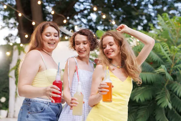 Üç genç kadın, açık hava yaz partisinde ışık ve kahkahalarla renkli şişe içeceklerin keyfini çıkarıyorlar..