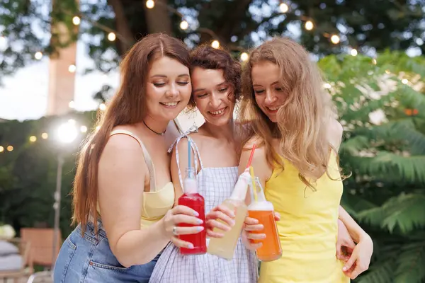 Üç genç kadın neşeli bir yaz partisinde renkli gazoz şişelerini tokuşturuyor, gülümsüyor ve neşeli havanın tadını çıkarıyorlar..
