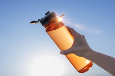 Mavi gökyüzüne karşı yeniden kullanılabilir bir su şişesini tutan bir kadının elinin yakın plan çekimi, susuzluk, sürdürülebilirlik ve açık hava yaşam tarzı kavramı.