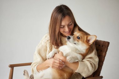 Gülümseyen kadın bir sandalyede sevimli bir Corgi köpeğine sarılıyor. Sıcak bir kış anı. Tarafsız bir arka planda dostluk ve aşk..