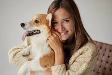 Gülümseyen kadın, mutlu Corgi köpeğine sarılıyor, samimi bir arkadaşlık ve şefkat anı, kapalı mekanda doğal ışıkla iç içe..