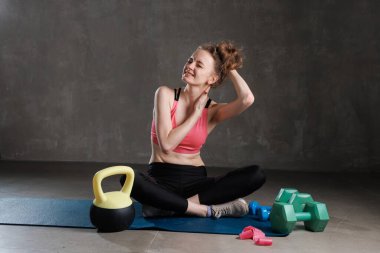 Spor kıyafetli genç bir kadın yoga minderinde oturuyor, antremandan sonra boyun ağrısı çekiyor, ağrıyan kaslara dokunuyor..