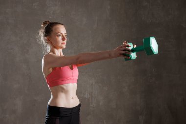 Odaklanmış fit bir kadın spor salonunda ağırlık çalışırken gri duvar arkasına doğru dambıl kaldırıyor..