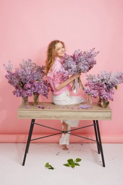 Pembe elbiseli neşeli kadın taze bahar çiçekleriyle çevrili bir masada otururken leylak buketi tutuyor..