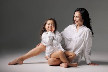 Neşeli anne-kız, büyük beyaz tişörtler giyip stüdyoda oturuyor, birlikte gülüyor ve kaynaşıyorlar..