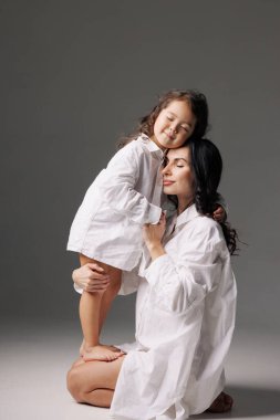 Sevgi dolu anne, çıplak ayakla kızını kucaklıyor. Büyük beyaz tişörtler içinde, gözleri kapalı, şefkatli bir aile anı..