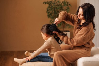 Genç anne, kızlarını sıcak ev ortamında uzun saçlarıyla tarıyor. Günlük bakım ve sevgi dolu bağlar kuruyor.