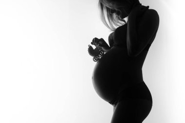 Bebek ayakkabısı tutan hamile bir kadının silüeti anneliği ve beklentiyi vurguluyor. Görüntü siyah beyaz, yumuşak ve duygusal bir atmosfer yaratıyor..