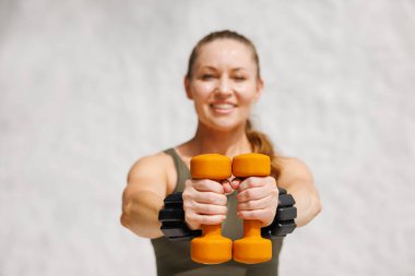 Formda bir kadın gülümsüyor ve elinde turuncu halterler tutuyordu. Spor yaparken motivasyon, güç ve sağlıklı yaşam tarzını gösteriyordu..