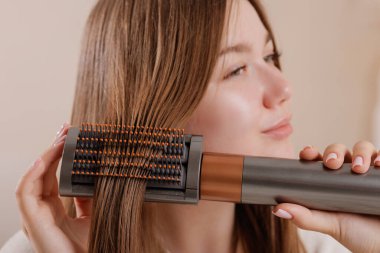 Evdeki uzun ve sağlıklı saçlarını düzleştirmek ve düzleştirmek için modern elektrikli saç tarama fırçası kullanan bir kadının yenilikçi bir güzelliği ve kendine özel bakım rutinini sergilediği yakın plan..