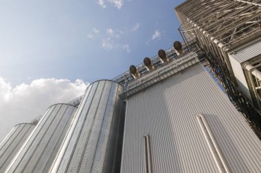 Mavi gökyüzü altındaki modern metal tahıl ambarları ve asansör tesisi, tarım endüstrisi ve depolamayı simgeliyor..