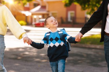 Sırt çantalı mutlu çocuk, anne ve babasının elini tutuyor, okula döndüğü ilk gün gülümsüyor, aile desteği, açık hava sabahı... 