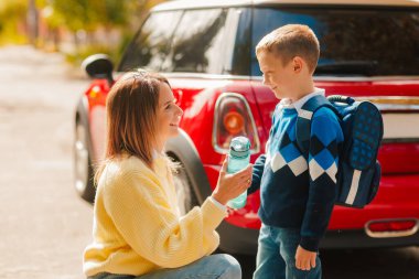 Gülümseyen anne, okula gitmeden önce sırt çantalı oğluna su şişesi verir.
